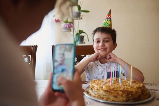 Cheerful adorable handsome Caucasian teenage boy sits at table with birthday cake with candles, smiles while posing to his blurred mom taking photo of him on smartphone in foreground. Birthday party