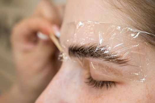 The master applies the composition for laminating the eyebrows to the client with a wooden spatula