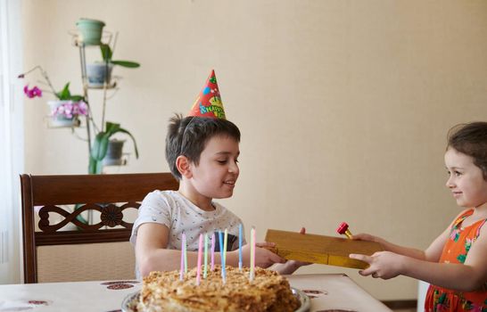 Charming cheerful European boy receives a gift from his younger sister for his 10th birthday, sits at the table with a birthday cake and candles. Birthday, anniversary and event celebration concept