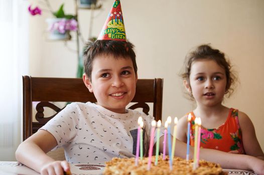 Cheerful handsome 10 year old boy sitting at the table with a delicious cake with lit birthday candles, next to his blurry little sister celebrating together birthday party