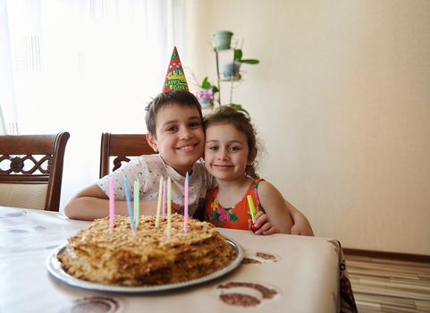 Portrait of two adorable cute Caucasian children, brother and sister, sitting at the table with a birthday cake decorated with long colorful candles. Anniversary and birthday party concept