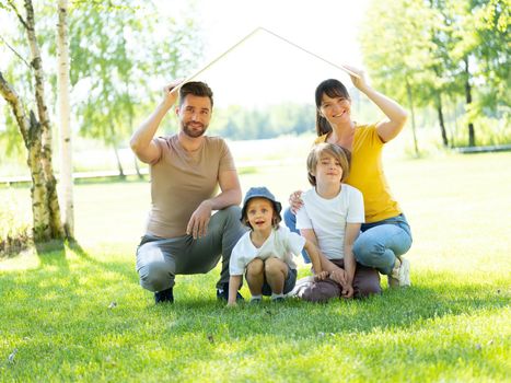 Concept of mortgage and housing for young families. Mother father and two children sitting on grass and holding roof symbol