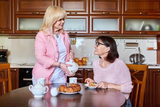 Two smiling happy mature women girlfriends in the kitchen together, having fun drinking tea chatting. Friendship, leisure, communication, 40s 50s age people concept