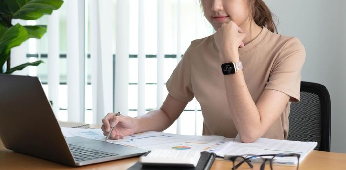 Beautiful Asian businesswoman analyzes charts using laptop calculator at the office..