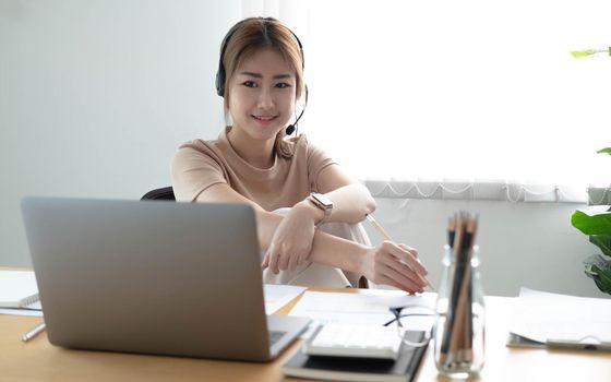 Asian businesswoman talking to colleague team in video call conference writing note on book with smile face. woman using computer laptop and headphone for online meeting.