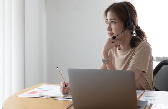 Asian businesswoman talking to colleague team in video call conference writing note on book with smile face. woman using computer laptop and headphone for online meeting.