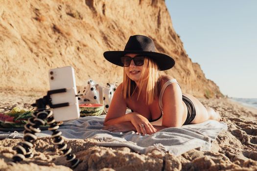 Young Woman in Sunglasses and Hat Lying on Beach, Filming Herself on Smartphone Attached on Tripod