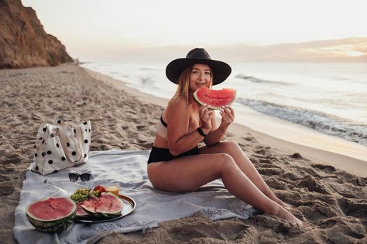 Young Pretty Woman Holding Slice of Watermelon While Sitting on the Plaid on Sandy Beach by Sea at Early Morning