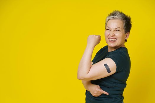 Senior grey haired woman rebelling showing nude shoulder with dark band aid or plaster on it isolated on yellow background. Amazing mature woman 50s. Vaccination and healthcare concept.