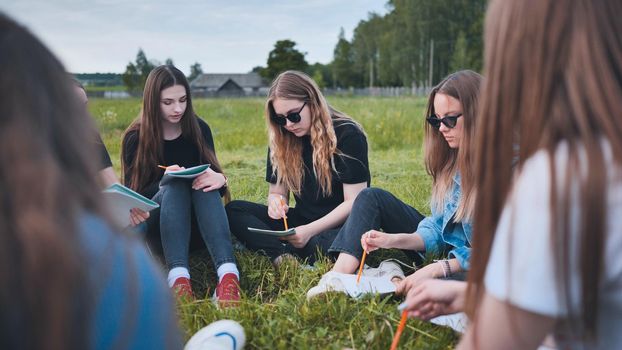 A group of female students are sitting in a circle on a meadow for collective work with notebooks