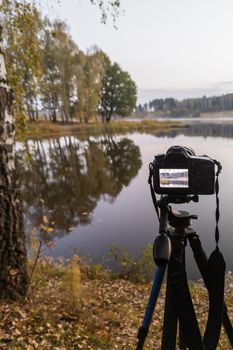 black digital camera on tripod shooting foggy early morning landscape at autumn lake with selective focus.