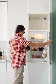 Delivery food, products to home. Shopping and healthy food concept. Young man in red plaid shirt holding disposable plastic boxed with food and putting them to the refridgerator