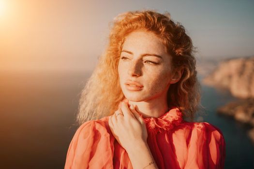 Side view a Young beautiful sensual woman in a red long dress posing on a volcanic rock high above the sea during sunset. Girl on the nature on overcast sky background. Fashion photo