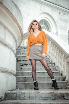 A middle-aged woman looks like a good blonde with curly beautiful hair and makeup on the background of the building. She is wearing a yellow sweater