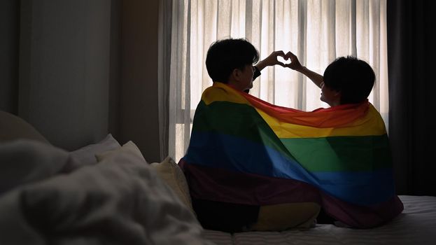 Happy same sex male couple making heart with their hands while sitting under rainbow flag. LGBT, pride, relationships and equality concept.