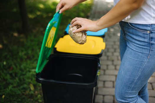 Closeup portrait woman hand throwing crumpled paper bag in recycling bin. Outdoors recycling bins different colours. Green cover.