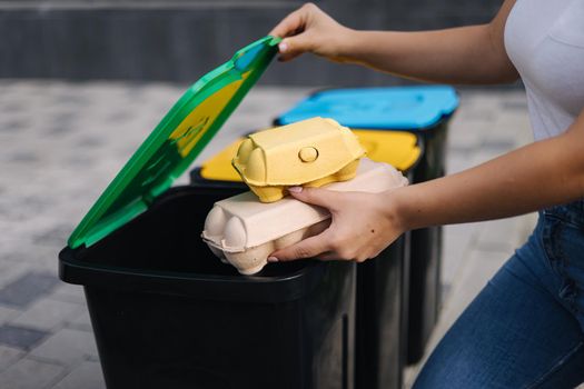 Middle selection of female hold two rubbish, plastic bottle and egg carton and thowing into different recycling bins. Close-up of woman stand by blue, yeallow and green bin.