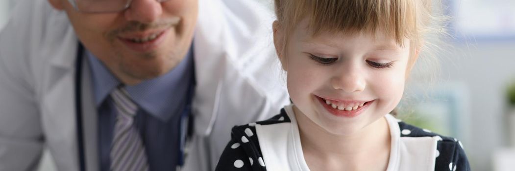 Portrait of little girl on pediatrician appointment in clinic, smiling kid get checkup from doctor. Medical worker in gown wearing stethoscope. Medicine, healthcare, childhood concept