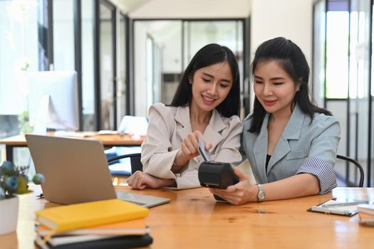 Two cheerful asian woman swiping a debit card through payment terminal at coffee shop.