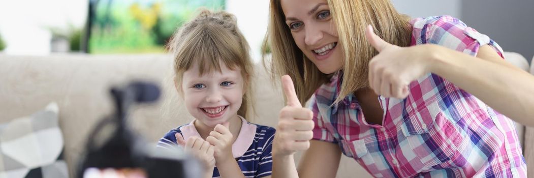 Portrait of mother and daughter showing thumbsup on video, videocamera set on tripod. Little girl and woman having fun together. Family time, childhood, pastime, holiday, friendship concept