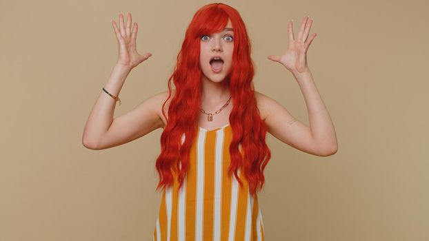 Oh my God, Wow. Excited amazed winner ginger woman raising hands in surprise looking at camera with big eyes, shocked by sudden victory. Sincere young girl isolated alone on beige studio background