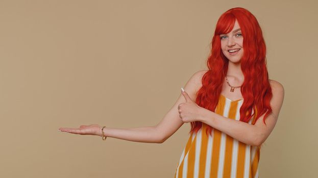 Beautiful redhead woman showing thumbs up, pointing empty place, advertising area for commercial text, copy space for goods promotion. Young ginger girl. Studio shot indoors on beige wall background