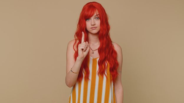 Displeased woman shakes finger and saying no, be careful, scolding and giving advice to avoid danger mistake, disapproval, disagree warning sign. Redhead girl isolated alone on beige studio background