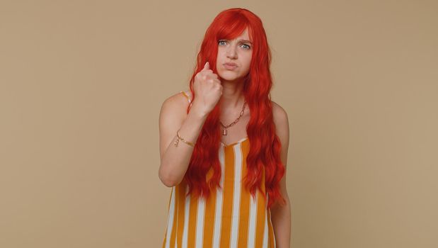 Aggressive angry ginger woman in tank top trying to fight at camera, shaking fist, boxing with expression, punishment, disappointment. Young redhead girl isolated alone on beige studio wall background