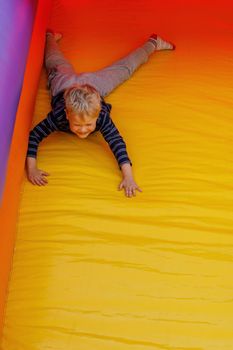 Little boy is actively playing on a children's inflatable trampoline. He slides down on his stomach. Bright red-yellow colors. Sunny day.