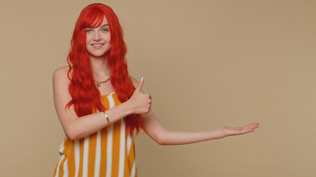 Beautiful redhead woman showing thumbs up and pointing empty place, advertising area for commercial text, copy space for goods promotion. Young ginger girl. Indoors studio shot on beige background