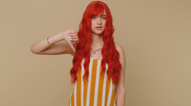 Upset unhappy red hair woman in striped tank top showing thumbs down sign gesture, expressing discontent, disapproval, dissatisfied, dislike. Young ginger girl. Indoor studio shot on beige background