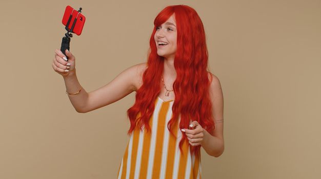 Redhead woman in tank top traveler blogger taking selfie on mobile phone selfie stick, communicating video call online with subscribers. Young ginger girl isolated alone on beige studio background
