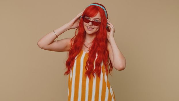 Portrait of redhead woman in tank top listening music via headphones and dancing disco fooling around having fun expressive gesticulating hands. Young one ginger girl on beige studio wall background