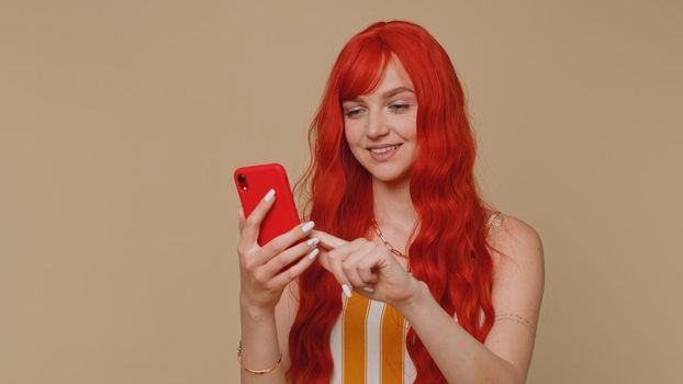 Beautiful red hair woman using mobile phone typing new post on web sms message, browsing, searching addiction of social networks. Young ginger girl studio shot isolated alone on beige wall background