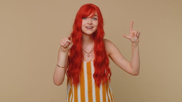 Woman showing loser gesture and pointing on you, blaming accusing for unsuccess, expressing disrespect, mocking your failure, bullying abuse. Young redhead girl isolated on beige studio background