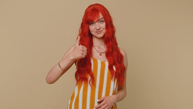 Redhead young woman in striped tank top raises thumbs up agrees with something or gives positive reply recommends advertisement likes good. Adult ginger girl isolated alone on beige studio background