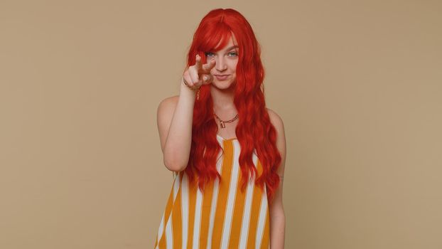 I am watching you. Ginger woman in tank top pointing at her eyes and camera, show I am watching you gesture, spying on someone. Young adult red hair girl isolated alone on beige studio wall background