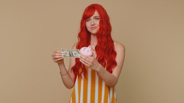 Poor woman upset over insufficient amount of money, holding piggybank and one dollar banknote. Financial crisis. Bankruptcy. Poverty and destitution. Young girl isolated on beige studio background
