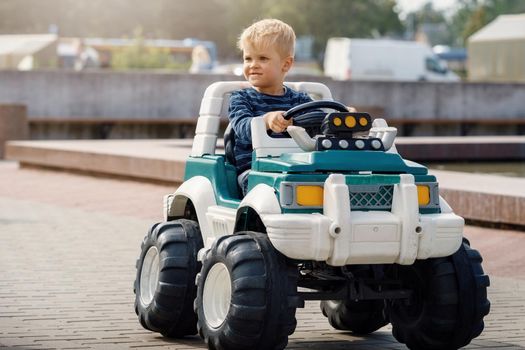 Cute little boy driving big electric toy car with steering wheel and having fun outdoors.