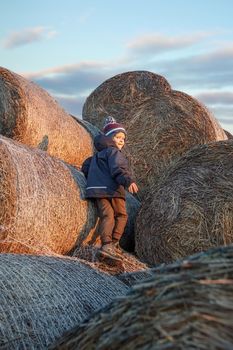 The little boy, wearing autumn clothes and a grandmother's knitted hat, plays outside on a pile of soft straw bales on an autumn evening.