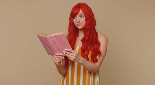 Redhead young woman reading funny interesting fairytale story book, leisure hobby, knowledge wisdom, education, learning, study, wow. Red hair ginger girl isolated alone on beige studio background