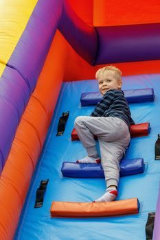 A cheerful smiling boy sports in the amusement park climbs the high stairs of an inflatable trampoline.
