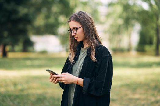 Young woman with glasses reading a message on her phone while standing in the park. High quality photo