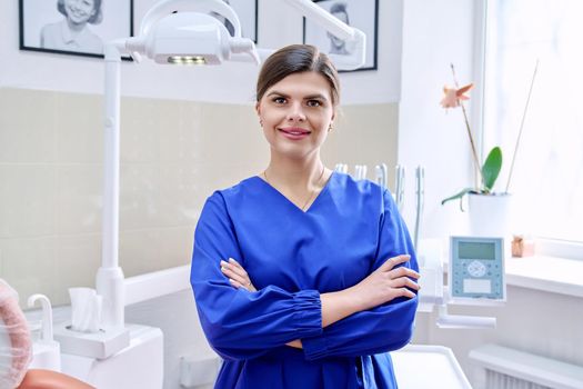 Portrait of confident female dentist in office looking at camera. Young female doctor in blue uniform with crossed arms. Dentistry, medicine, health care, profession, stomatology concept