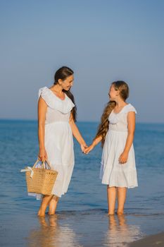 mom and daughter in white dresses walk on the beach