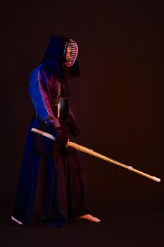 Masculine Kendo master wearing in an armor, traditional kimono and helmet is practicing martial art with the shinai bamboo sword posing standing sideways against a black studio background. Combat training, sportsmanship, close up. Full length, barefoot.