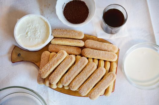 Flat lay composition of Tiramisu ingredients: savoiardi biscuit cookies on a wooden board and a bowl with mascarpone cheese for The Italian dessert Tiramisu. Copy ad space.