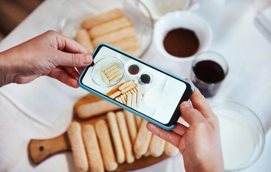 View from above. Hands using smartphone camera and taking photo of ingredients for Tiramisu dessert on the white tablecloth background. Mobile phone in live view mode