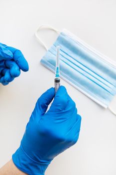 A hand in a blue glove holding a syringe with a medicine against the background of a medical mask. Vaccination and health concept