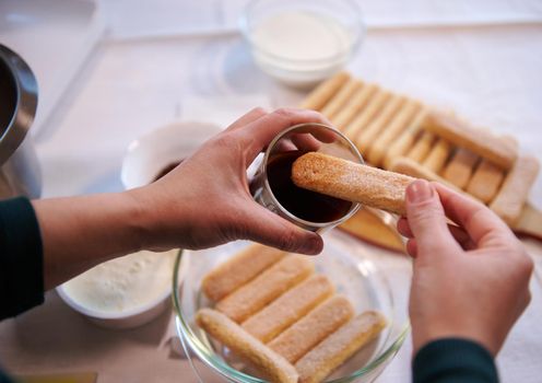 Details of a confectioner hands dipping ladyfinger biscuit cookies into a glass of strong sweet espresso coffee. Step-by-step recipe for making traditional Italian dessert Tiramisu at home. Culinary